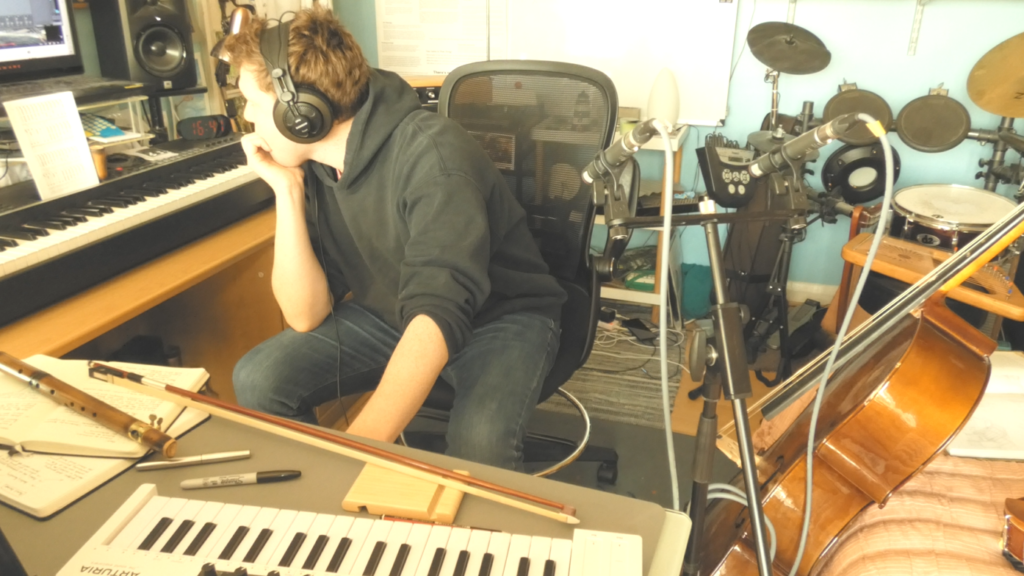 Screenshot 2025-06-21 194851 Felix sitting in his studio looking at computer. On desk is a bamboosax, violin bow and two electric piano keyboards.